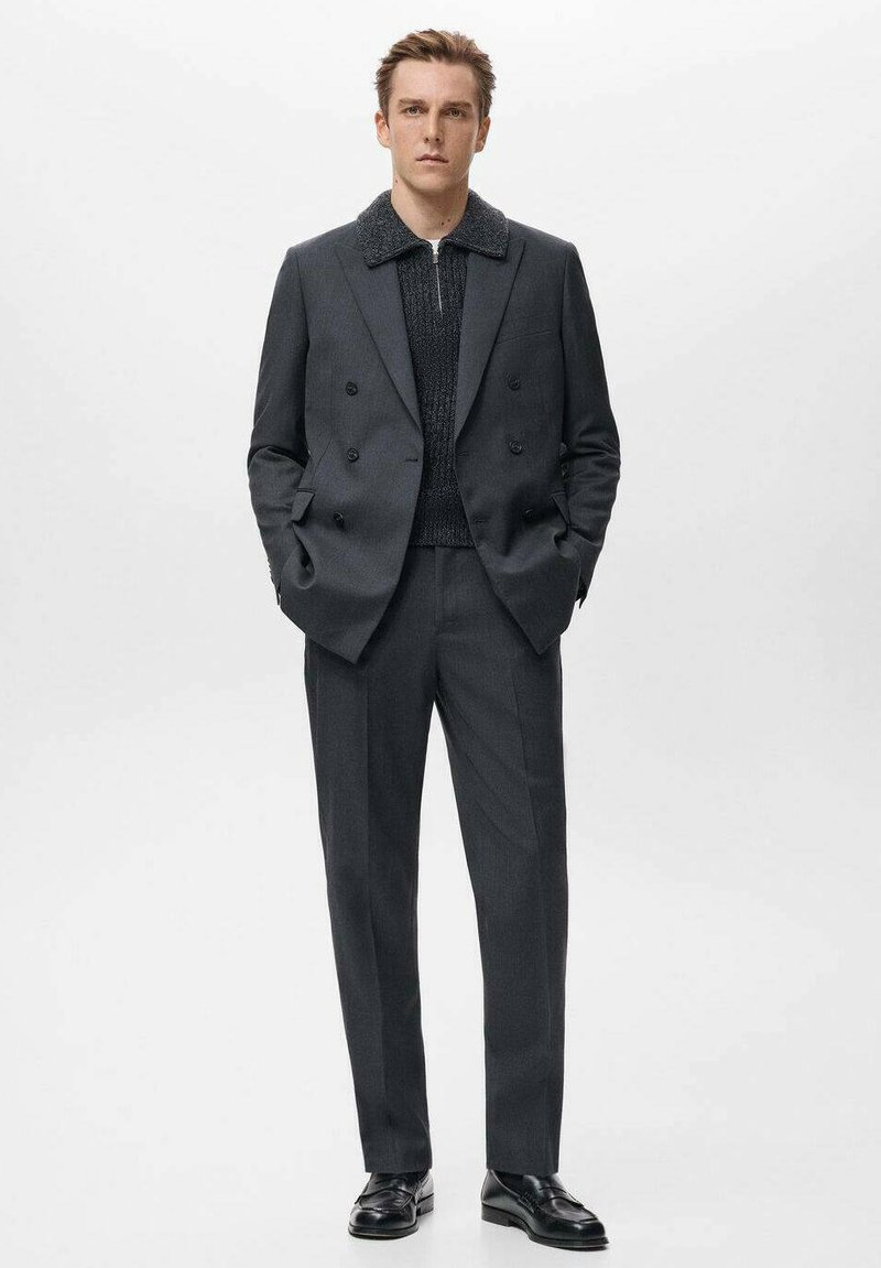 Abito doppiopetto grigio con camicia a maglia scura testurizzata e ampio colletto, abbinato a scarpe nere lucide, in piedi contro uno sfondo bianco.