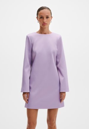 Kvinne iført en lilla minikjole med lange ermer, stående foran en enkel hvit bakgrunn, og ser rett mot kameraet.