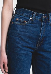 Dark blue denim jeans feature a high waist, five-pocket design, copper hardware accents, and contrasting orange stitching. Soft texture visible.