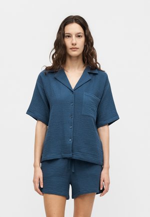 Vrouw met lang golvend bruin haar, die een blauw kort mouw knoophemd met borstzak en bijpassende shorts draagt tegen een effen achtergrond.