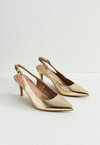 Un par de tacones slingback metálicos dorados con puntas afiladas y correas con hebilla sobre una superficie blanca y un fondo liso.
