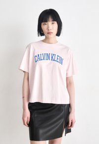 Rozā kokvilnas t-krekls ar zilu "CALVIN KLEIN" druku, apvienots ar melnu ādas svārku ar sānu šķēlumu, gludu tekstūru un cieši pieguļošu dizainu.
