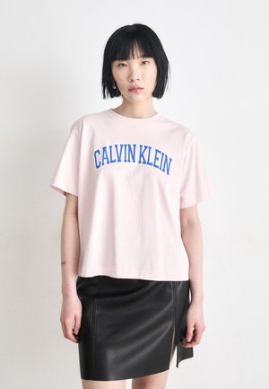 Rozā kokvilnas t-krekls ar zilu "CALVIN KLEIN" druku, apvienots ar melnu ādas svārku ar sānu šķēlumu, gludu tekstūru un cieši pieguļošu dizainu.