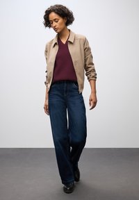 Beige Bomberjacke mit elastischen Bündchen, bordeauxroter V-Ausschnitt-Pullover, dunkelblaue weit geschnittene Jeans und schwarze Schuhe vor einem neutralen Hintergrund.