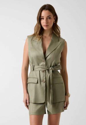 Mujer con blazer sin mangas color oliva con cinturón y bolsillos delanteros, y pantalones cortos a juego, de pie contra un fondo claro y liso.