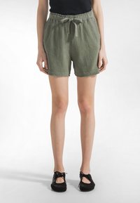 Frau trägt olivgrüne, strukturierte Shorts mit Kordelzug in der Taille und schwarze Schnürschuhe, steht vor einem weißen Hintergrund.