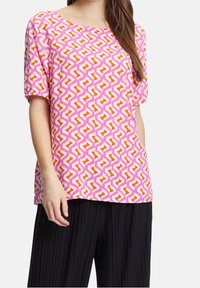 T-shirt à manches courtes rose avec un motif géométrique de zigzags orange et blanc. Tissu léger avec une coupe décontractée. Associé à un pantalon noir plissé.