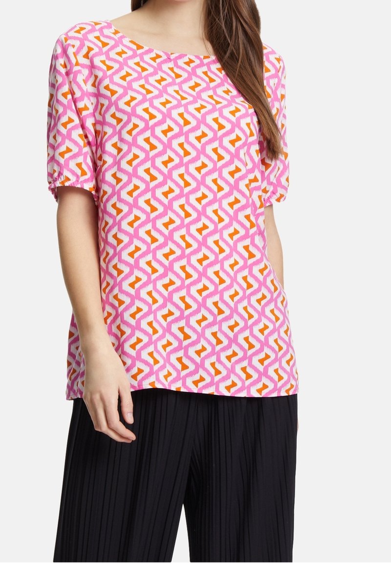 T-shirt à manches courtes rose avec un motif géométrique de zigzags orange et blanc. Tissu léger avec une coupe décontractée. Associé à un pantalon noir plissé.