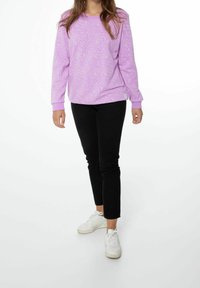 Felpa viola con macchie bianche, polsini a costine, abbinata a pantaloni neri slim-fit e sneakers bianche, in piedi su uno sfondo bianco.