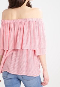 Blusa rosa a righe, scollata sulle spalle, con strati arricciati e maniche corte; tessuto leggero con vestibilità rilassata. Indossata con jeans blu.
