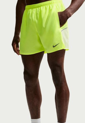 Mężczyzna w jaskrawożółtych sportowych szortach Nike z elastycznym pasem i boczną kieszenią, stojący z jedną ręką w kieszeni i drugą zrelaksowaną.