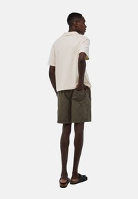 Polo shirt beige à manches courtes, coupe décontractée, associé à un short vert olive. Le modèle porte des sandales noires et adopte une posture neutre.