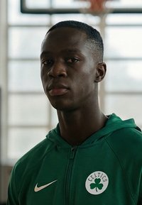 Noor mees rohelises Celticsi kapuutsiga, kes seisab toas, mille taustal on uduse kehaga korvpallikaar.