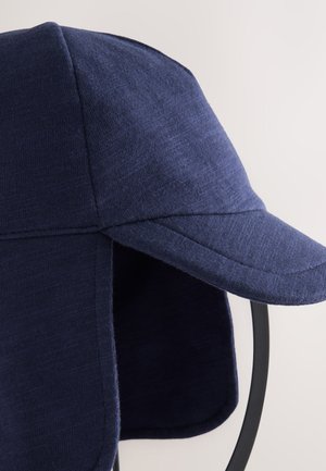 Tamsiai mėlyna kepurė, pagaminta iš minkštos medžiagos, turi išlenktą snapelį ir ausų uždangalus papildomam apsaugai. Lygus tekstūra su minimaliais siuvimo elementais.