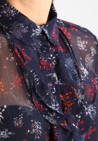 Blusa floreale navy con un materiale trasparente, caratterizzata da motivi di fiori rossi e bianchi, un colletto classico e chiusura con bottoni al collo.