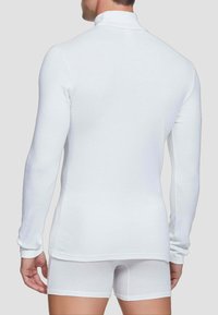 Maglia bianca a maniche lunghe con colletto alto, realizzata in tessuto morbido. La vista dal retro mostra un design aderente e un orlo leggermente più lungo.