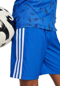Blauw voetbalshirt met een geometrisch patroon en blauwe shorts met drie witte strepen langs de zijkanten, terwijl hij een zwart-wit voetbal vasthoudt.