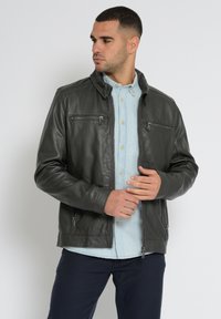 Donkergrijze leren jas voor mannen met ritszakken en een staande kraag, gelaagd over een lichtblauw overhemd en donkere broek. Gladde textuur.