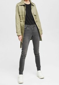 Jasnooliwkowy płaszcz do kostek z zapięciem na guziki i dwoma kieszeniami, w połączeniu z czarną koszulą, szarymi zwężanymi dżinsami i białymi trampkami.