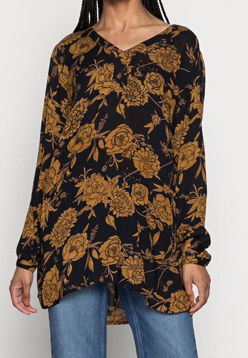 Femme portant un chemisier noir ample à manches longues avec un motif floral jaune moutarde et un jean bleu, debout les bras détendus.