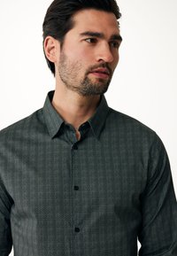 Camicia button-up a motivo scuro con una trama a spina di pesce, bottoni neri e colletto strutturato. Il tessuto appare liscio e leggero.
