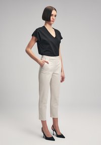 Haut noir à col en V et manches courtes, pantalon beige slim, escarpins noirs à bout pointu. Tissu lisse, coupe ajustée, accents en dentelle discrets sur les manches.