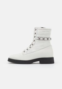 Bottine en cuir blanc avec un bout arrondi, fermeture à lacets à l'avant et accent en chaîne argentée sur le côté. Semelle en caoutchouc noire avec un petit talon.