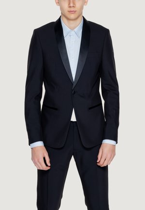 Muž v černém obleku na míru s hedvábnými límci, oblečený přes světle modrou košili s knoflíky, stojící před jednobarevným pozadím.