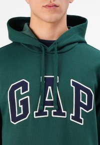 Green hoodie with a front pocket, featuring large white and navy "GAP" lettering. Soft fabric, adjustable drawstring hood, and ribbed cuffs.