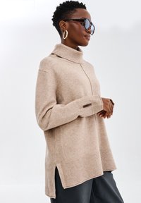 Beżowy sweter z golfem z prążkowanymi akcentami, z bocznymi rozcięciami i dopasowanym krojem. Model nosi ciemne spodnie i okulary przeciwsłoneczne. Widoczna miękka tekstura.