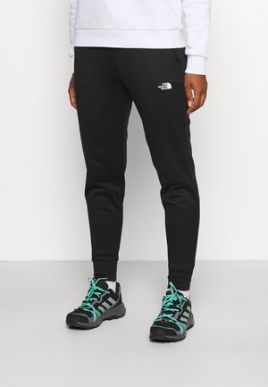 Osoba w czarnych, zwężanych spodniach dresowych The North Face i czarnych butach sportowych Adidas z turkusowymi sznurówkami, stojąca na jasnej podłodze.