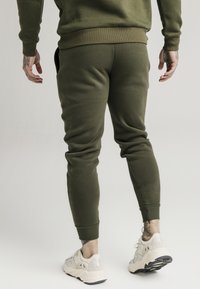 Olivgröna joggers i ett mjukt tyg, med ribbade mudd och sidofickor, i kombination med krämfärgade sneakers med nätinlägg och svart sula.