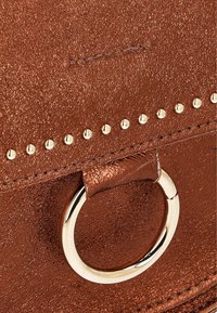Metallic brun håndtaske detaljer med en struktureret overflade, guldnagle accenter og en cirkulær guld ring-beslag.