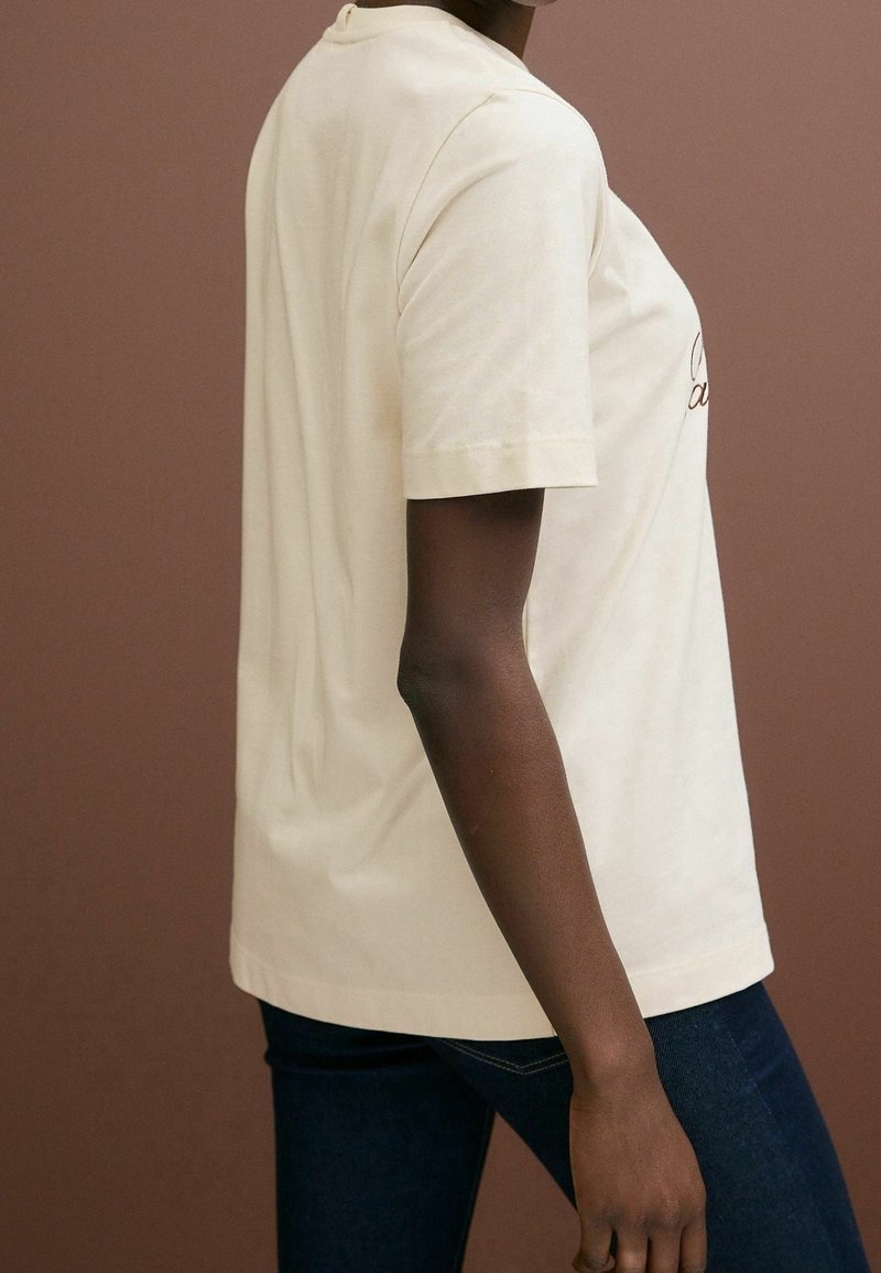 T-shirt en coton crème à manches courtes, coupe décontractée et encolure arrondie, avec texte brodé sur le devant.