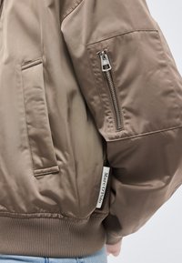 Mâneca unei jachete bomber taupe cu buzunar cu fermoar și buzunar lateral, purtată de o persoană în blugi albaștri.