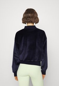 Pull en velours bleu marine avec un col haut, des épaules tombantes et des poignets côtelés, assorti à des leggings vert clair, mettant en valeur une texture douce.