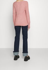 Osoba ubrana w różowy sweter z długim rękawem, ciemne dżinsy z mankietami i czarne masywne buty, stojąca na jednolitym jasnym tle.