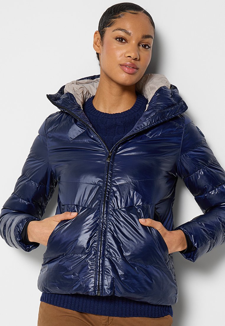 Dunkelblaues Steppjacke mit glänzender Oberfläche, Kapuze und Reißverschluss vorne. Verfügt über horizontale Nähte und zwei Seitentaschen, getragen über einem gestrickten Pullover.