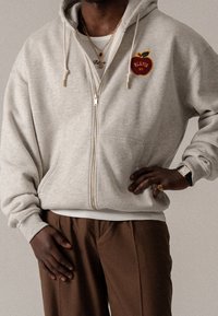 Jasnoszara bluza z kapturem zapinana na zamek, z przednią kieszenią, haftowanym logo jabłka i napisem, z prążkowanymi mankietami i dołem, noszona na brązowych spodniach.