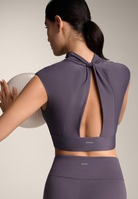 Top sportivo viola a maniche corte con colletto alto, apertura sulla schiena e dettaglio annodato. Tessuto morbido e elasticizzato, abbinato a leggings coordinati.