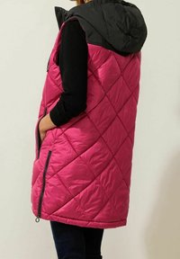 Rosa vadderad väst med svart huva, med diagonala sömmar, två sidofickor med dragkedja och en längre nederdel bak. Tillverkad av glänsande, lätt material.
