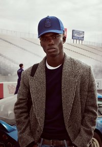 Das Modell trägt ein graues Blazer im Hahnentrittmuster über einem marineblauen Pullover, dazu eine blaue Kappe mit einem Logo. Der Hintergrund zeigt Rennwagen und ein Stadion.