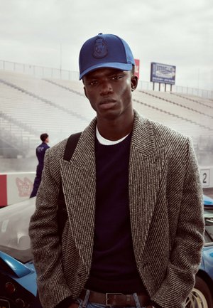 Le modèle porte un blazer à motif pied-de-poule gris par-dessus un pull bleu marine, ainsi qu'une casquette bleue avec un logo. L'arrière-plan comprend des voitures de course et un stade.