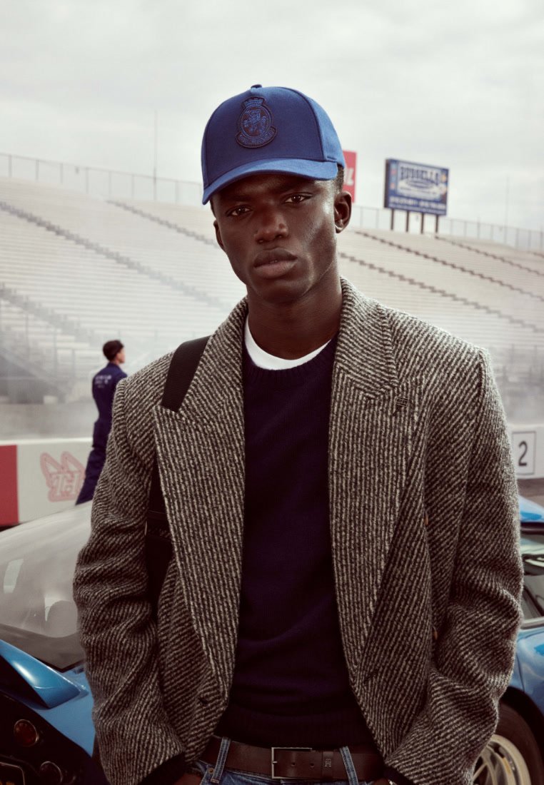 Das Modell trägt ein graues Blazer im Hahnentrittmuster über einem marineblauen Pullover, dazu eine blaue Kappe mit einem Logo. Der Hintergrund zeigt Rennwagen und ein Stadion.