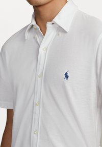 Polo bianco a maniche corte in tessuto strutturato, con logo ricamato blu, chiusura frontale a bottoni e design del colletto classico.