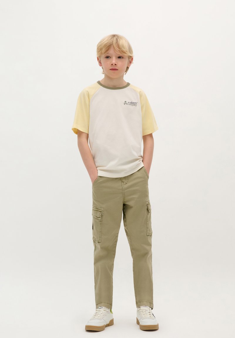 Witte en gele raglan T-shirt met groene accenten, gecombineerd met licht kaki cargobroeken met zijzakken. Witte sneakers met tan zolen.