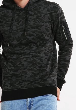 Mężczyzna noszący ciemnoszarą i czarną bluzę z kapturem w kamuflażu, z kieszenią na suwak na lewym rękawie, oraz jasnoniebieskie dżinsy.
