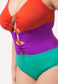 Maillot de bain une pièce coloré avec un haut rouge, une partie médiane violette avec des fronces et un bas vert. Les matériaux sont lisses avec une finition mate.