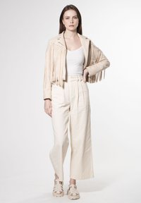 Giacca in suede beige con frange e dettagli in argento, abbinata a pantaloni a gamba larga crema e sandali gladiatore color nocciola. Texture morbide ovunque.