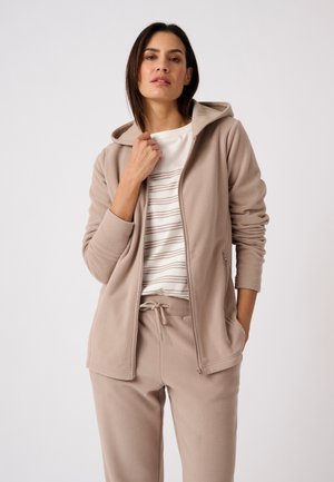 Donna che indossa una giacca beige con cappuccio e zip e pantaloni abbinati con camicia bianca a righe, posa con la mano vicino al colletto su sfondo semplice.